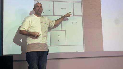 En man står på en scen och pekar på en presentation som projiceras bakom honom.