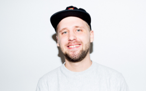 Marcus står mot en vit vägg och är ljust klädd med en svart keps på sig och ler stort.