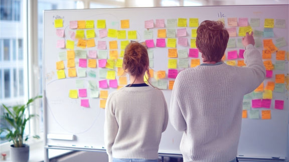 Två personer står framför en whiteboard täckt med färgglada post-it-lappar och arbetar med idé- och planeringsarbete.