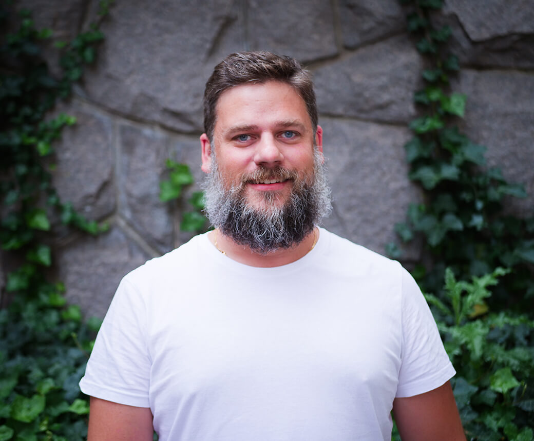 Daniel ler ståendes framför en stenmur med murgröna. Han har ett stort skägg och en vit t-shirt.