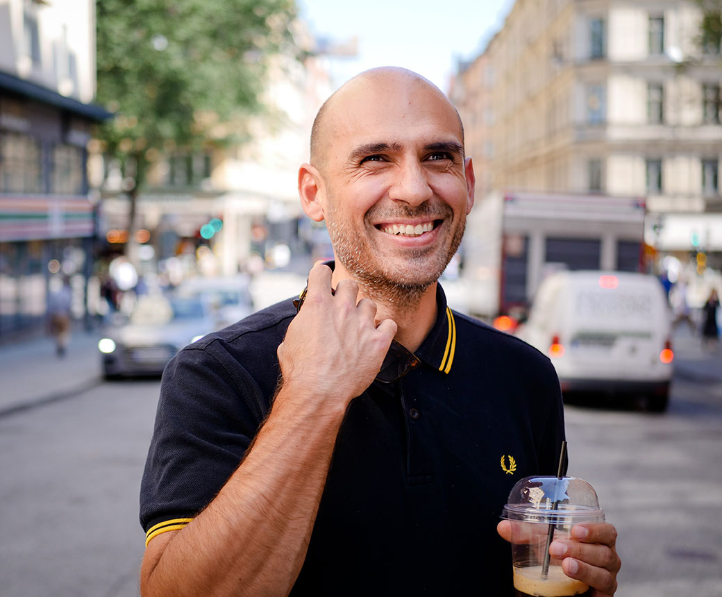 Dimitris har ett brett leende och tittar på något utanför bild. Han har mörk kortärmad tröja och håller i en takeaway-kaffe. I bakgrunden syns en trafikerad gata.