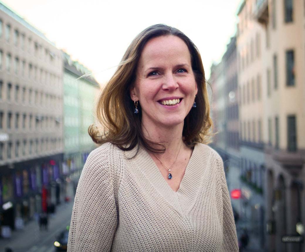 Karin ler ståendes uppe på en bro. Hon har axellångt mörkt hår och bär en beige stickad tröja. I bakgrunden syns en trafikerad gata.