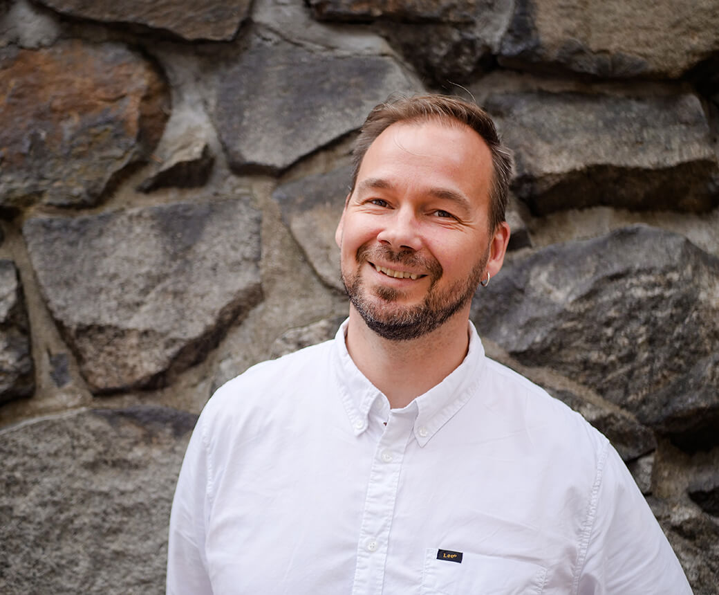 Mikael står utomhus vid en stenvägg. Han ler, har kortklippt skägg och bär vit skjorta.