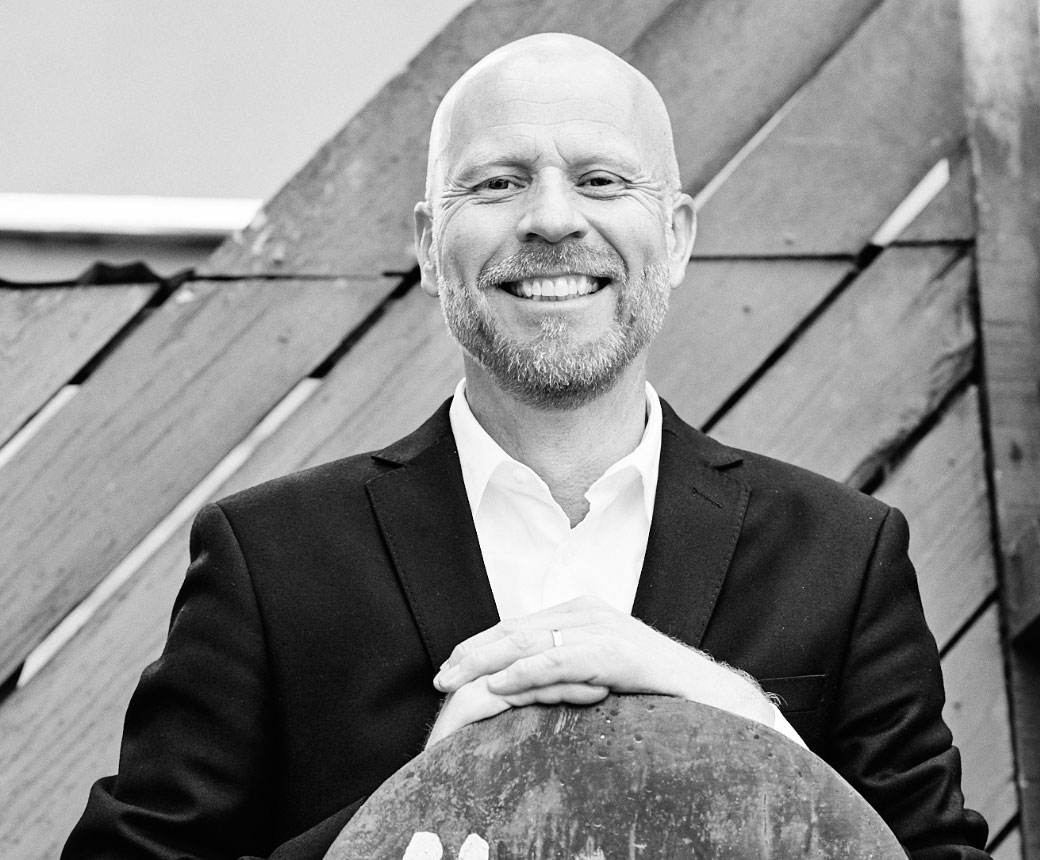 Per tittar rakt in i kameran och ler brett. Han har kort skäggstubb och en välsittande svart kostym. Svart-vitt foto.