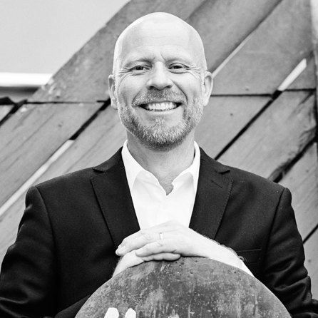 Per tittar rakt in i kameran och ler brett. Han har kort skäggstubb och en välsittande svart kostym. Svart-vitt foto.