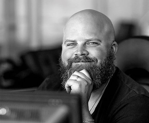 Putte sitter vid sitt skrivbord. Han ler självsäkert och håller upp en hand mot hakan. Hans skägg är stort och lockigt. Svart-vitt foto.