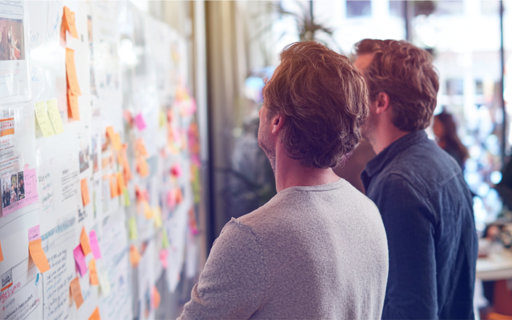 Två personer studerar en vägg fylld med utskrifter och färgglada post-it-lappar under ett digitalt strategiarbete.