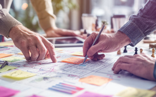 Närbild på händer som pekar och skriver på en planeringskarta fylld med post-it-lappar och diagram.