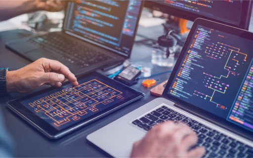 Personer arbetar vid datorer och surfplatta med tekniska systemkartor och kod på skärmarna.
