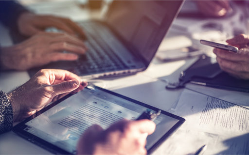 Händer håller en surfplatta med textdokument, medan andra arbetar på dator och mobil.