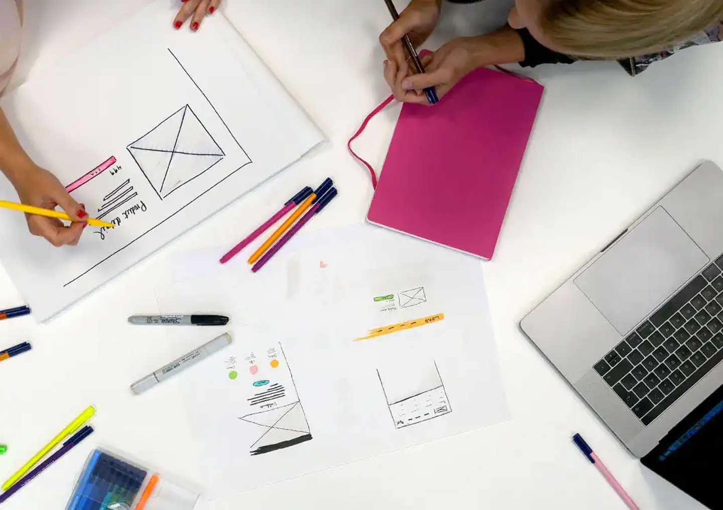 Två personer arbetar med UI-designskisser vid ett bord med färgpennor, papper med wireframes och en laptop.