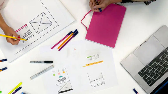 Två personer arbetar med UI-designskisser vid ett bord med färgpennor, papper med wireframes och en laptop.