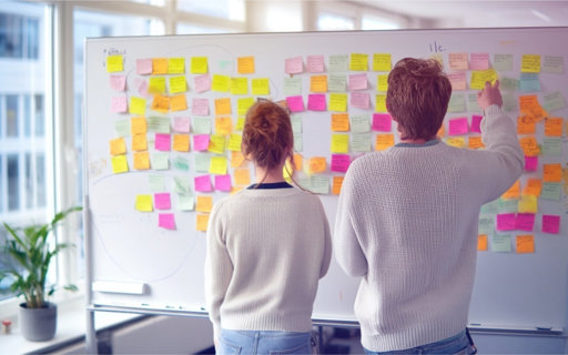 Två personer står framför en whiteboard täckt med färgglada post-it-lappar och arbetar med idé- och planeringsarbete.